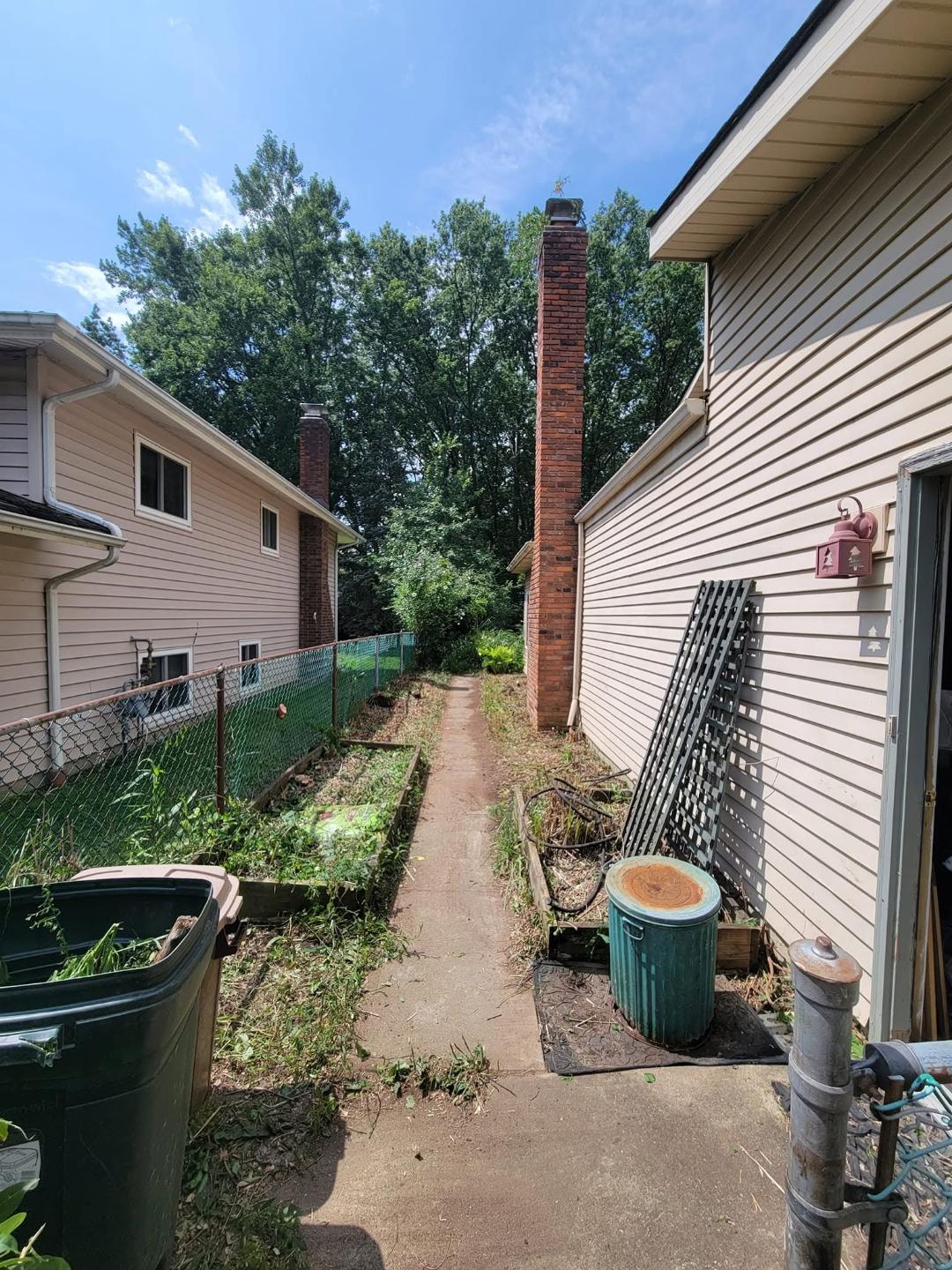 After photo of brush removal: cleared gravel path with fence, garden bed, trash bin, and clean siding beside beige house.