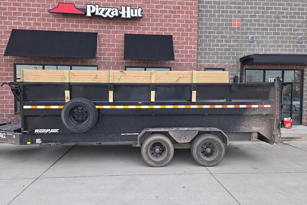 Black dual-axle dump trailer loaded with wooden pallets parked in front of a Pizza Hut restaurant building.