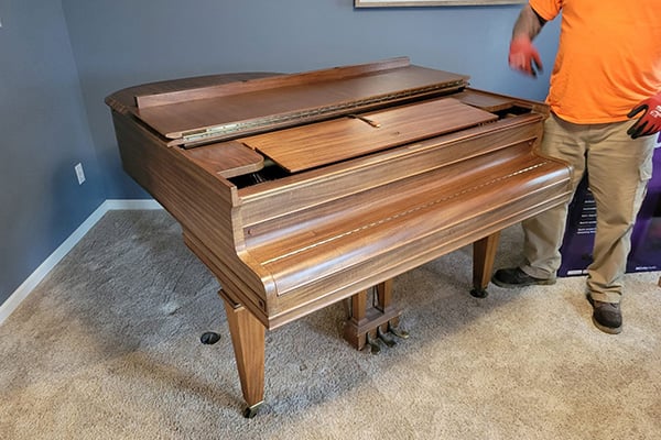 Worker in orange shirt dismantling wooden grand piano inside home with open lid and exposed internals.