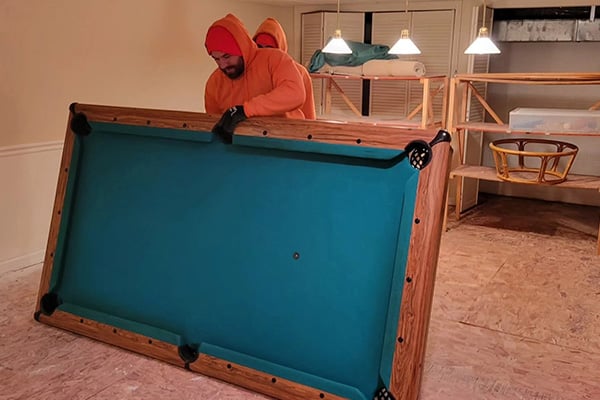 Workers in orange hoodies lifting wooden pool table with green felt from home interior during junk removal.