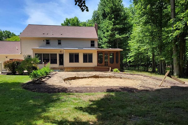 After photo of swimming pool removal: excavated empty pit beside beige house with clean yard, remaining deck, sunroom, and trees.