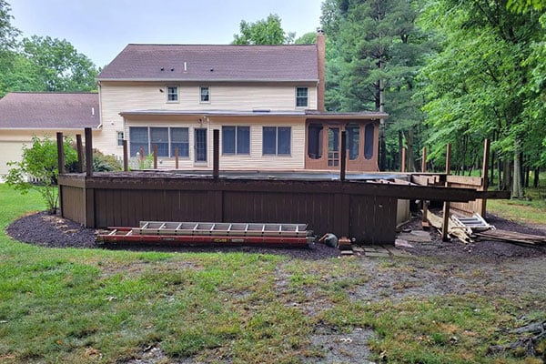 Before photo of swimming pool removal: partially demolished wooden deck beside beige house with lumber debris and trees.